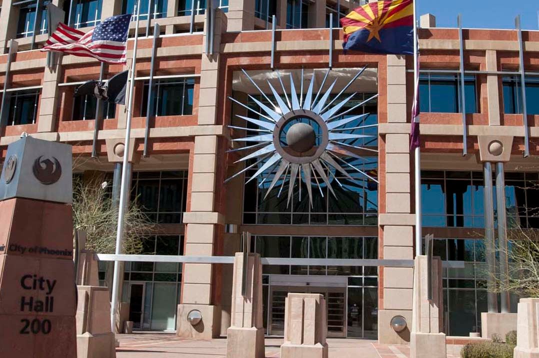 A photo of the front entrance to Phoenix city hall, including the U.S. and state flags, and a large sunburst sculpture that hangs over the door.