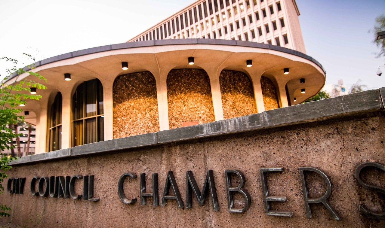 Photograph of City Council chambers. The image shows a low sign in the foreground, a round building in the midground, and a tall office building in the background.