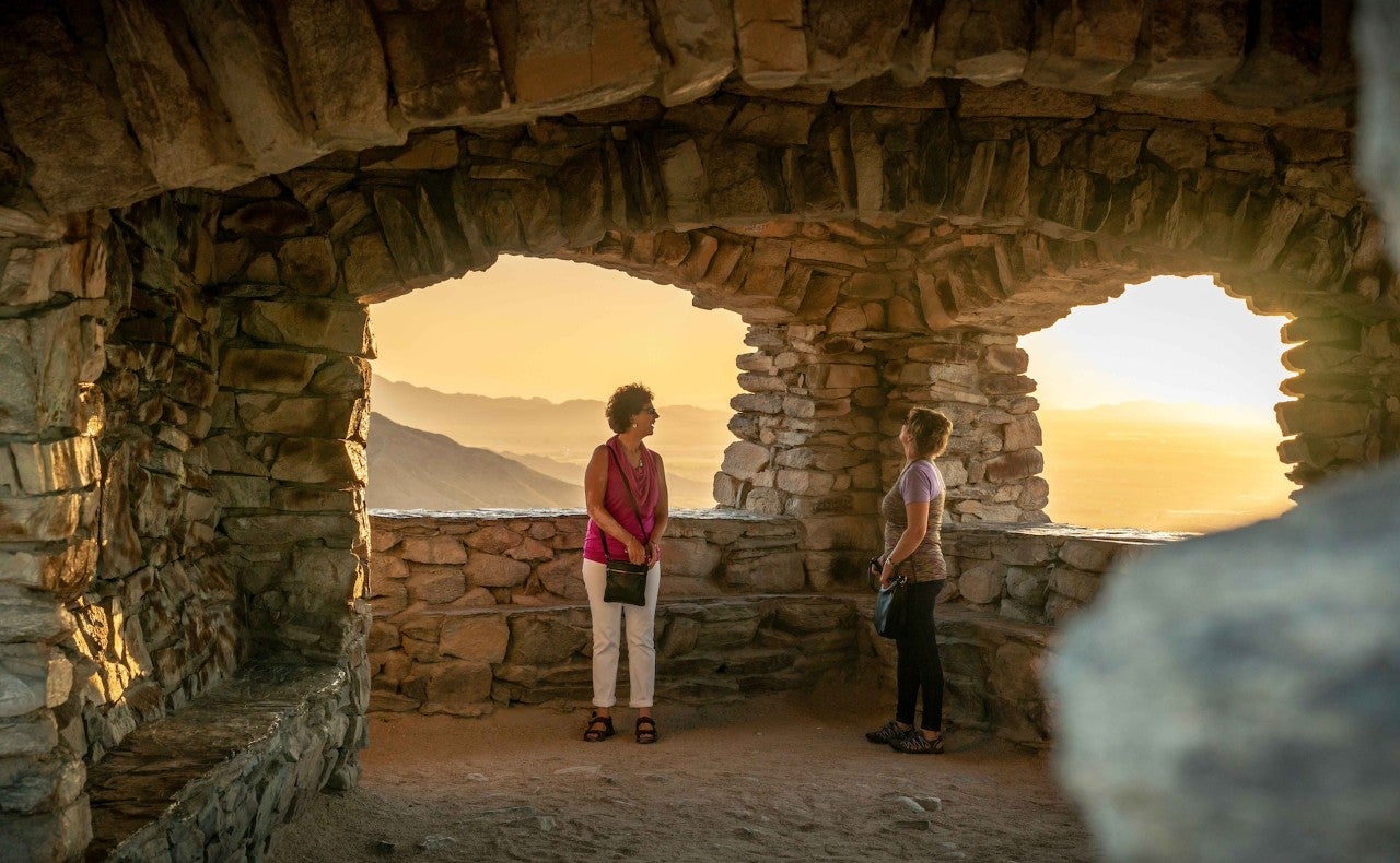 Two women stand in a stone structure on a mountain top during sunset.
